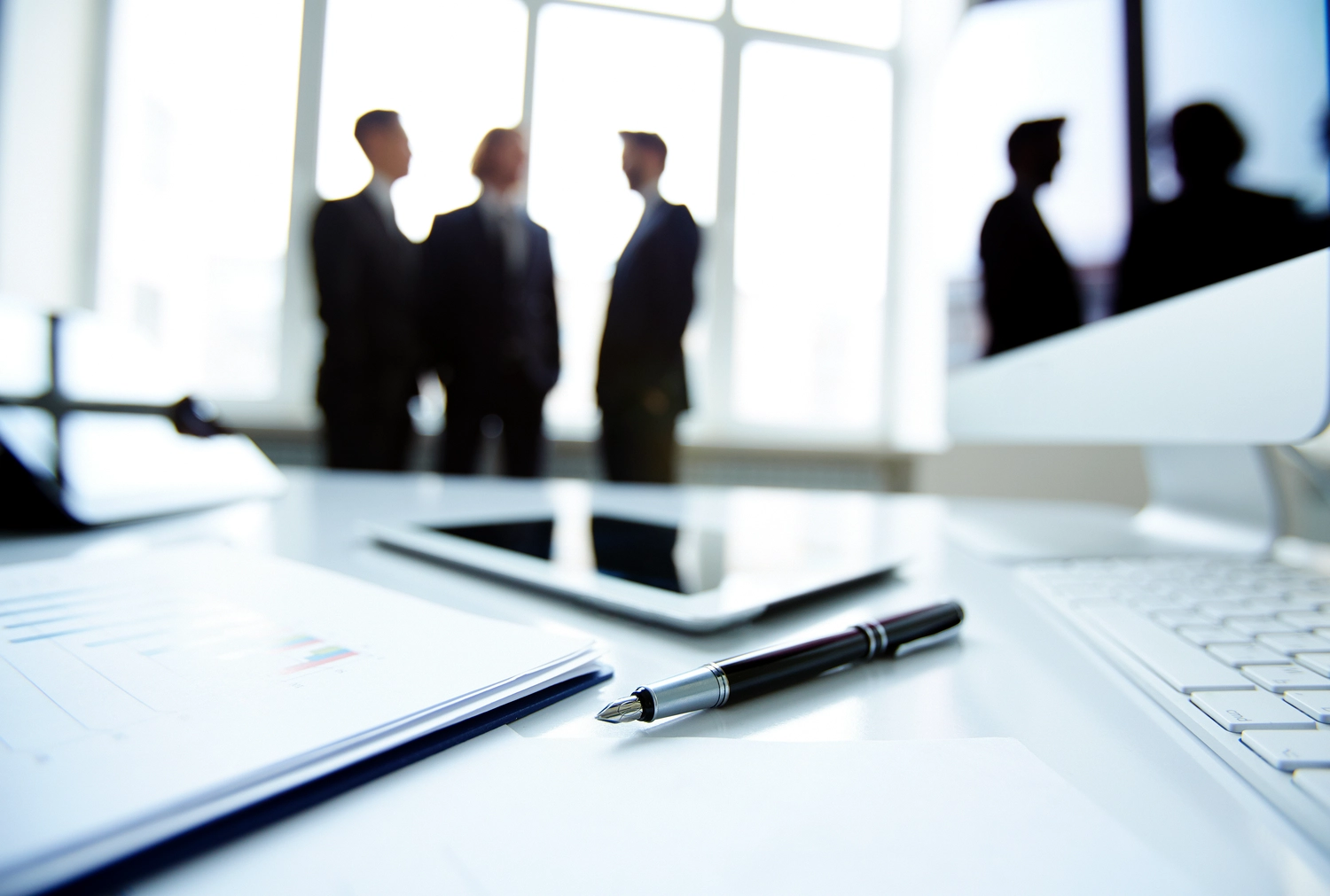 Mes West GmbH - Fachkräfte-, Investitions- und Unternehmenslösungen - Silhouetten von Geschäftsleuten in einem hellen, modernen Büro, gesehen durch ein Fenster. Im Vordergrund sind ein Schreibtisch und ein Tablet verschwommen zu erkennen.