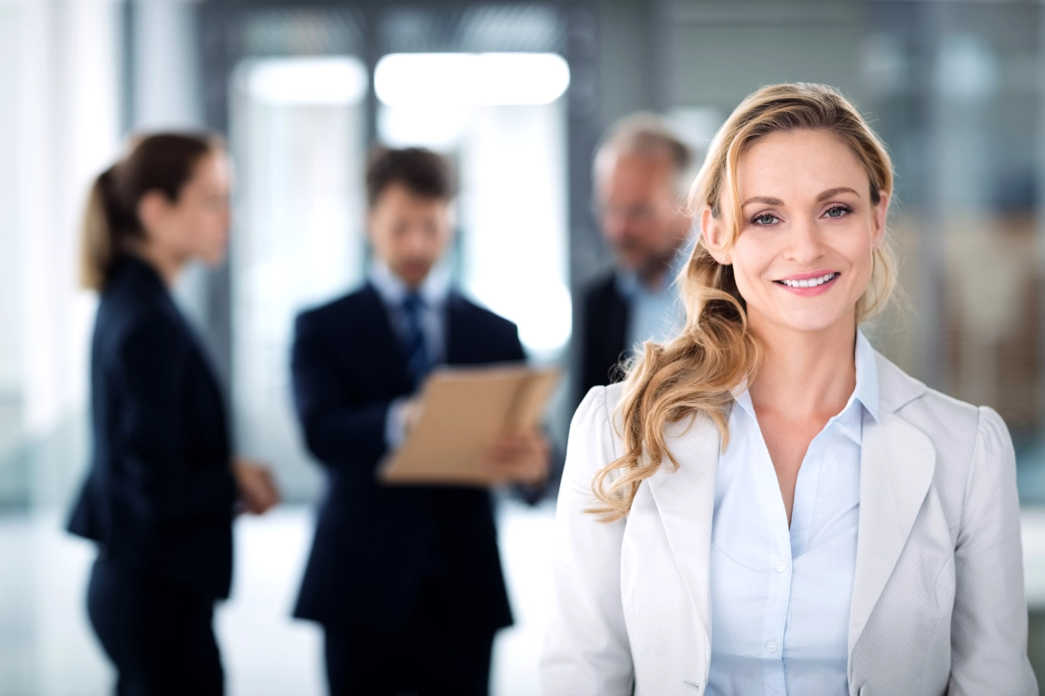 Mes West GmbH - Fachkräfte-, Investitions- und Unternehmenslösungen - Eine lächelnde blonde Frau in weißem Blazer und hellblauer Bluse steht in einem professionellen Büro. Im Hintergrund sind ihre Kollegen verschwommen zu sehen.
