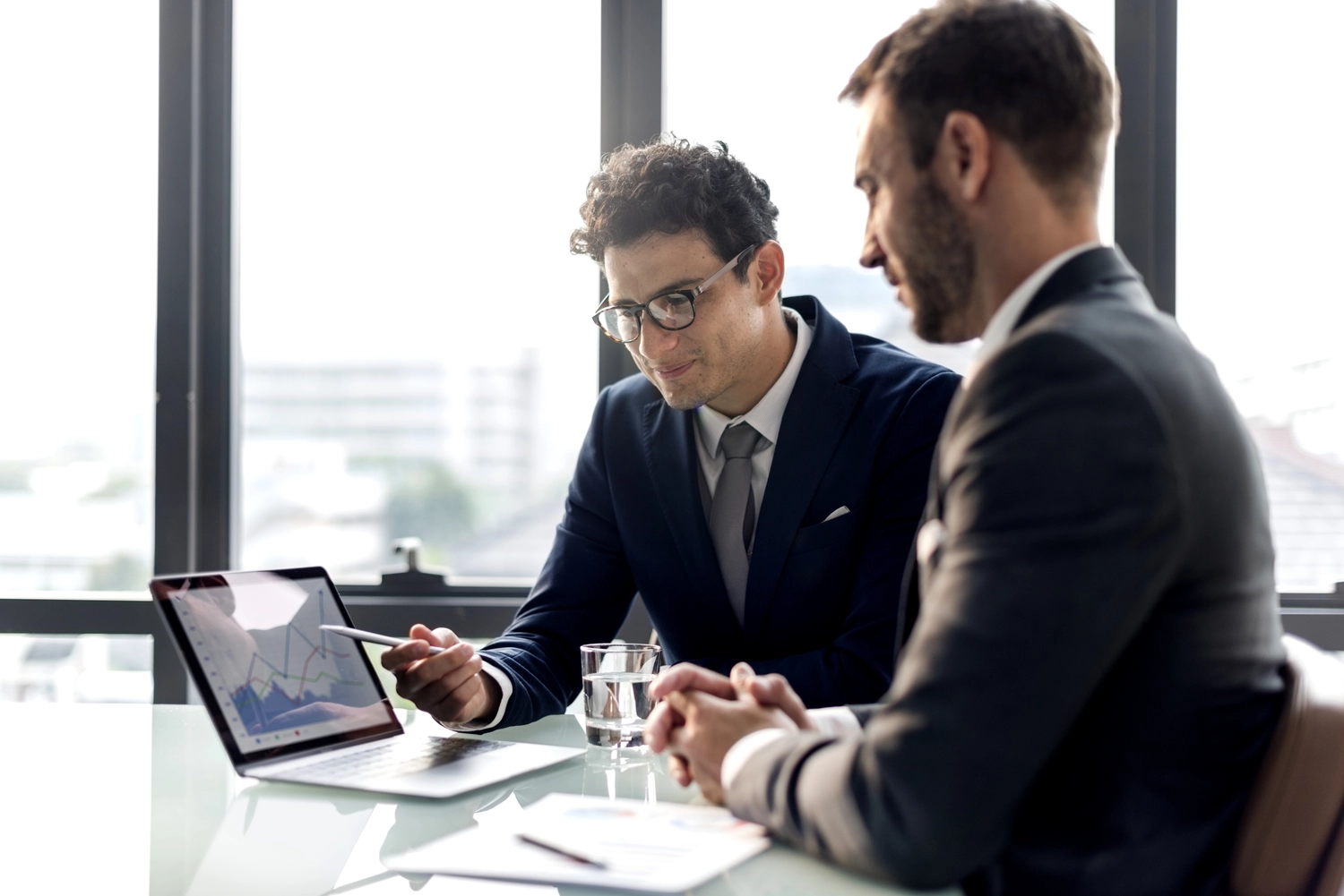 Mes West GmbH - Fachkräfte-, Investitions- und Unternehmenslösungen - Zwei Männer in professioneller Geschäftskleidung blicken in einem modernen Büro mit einem großen Fenster im Hintergrund auf ein Dokument oder Tablet.