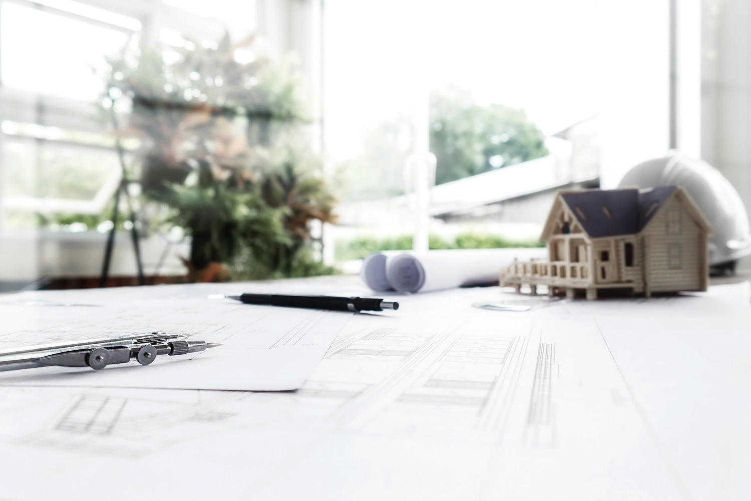 Mes West GmbH - Fachkräfte-, Investitions- und Unternehmenslösungen - Ein Architekturarbeitsplatz mit Bauplänen, einem Kompass und einem kleinen Holzhausmodell