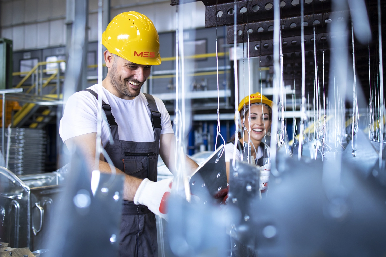 Mes West GmbH - Fachkräfte-, Investitions- und Unternehmenslösungen - Zwei Industriearbeiter, ein Mann und eine Frau, die gelbe Schutzhelme und Schutzkleidung tragen, lächeln bei der Arbeit in einer Produktionshalle, in der Metallteile von der Decke hängen.